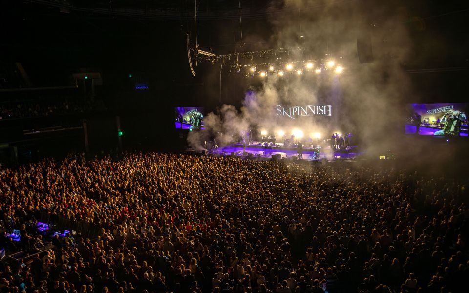 A packed crowd of people watch Skipinnish perform on stage, accompanied by spotlights and haze.