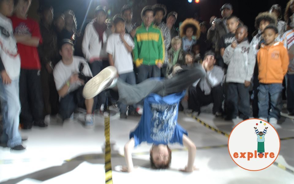 Blurry handheld image of a young person breakdancing, watched by a crowd of young onlookers. In the bottom right corner, a roundel featuring a playful illustration of a person juggling stood on the word 'explore'.