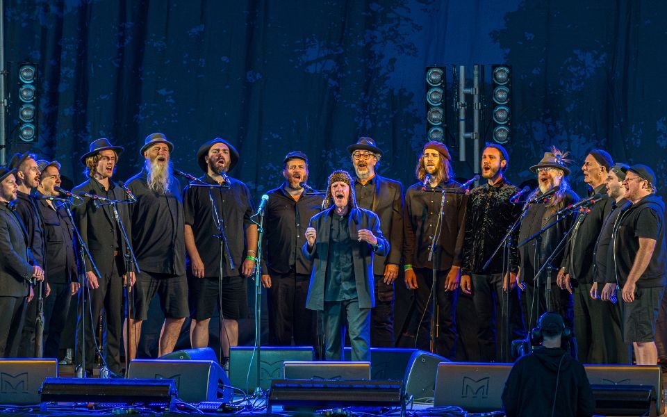 The Spooky Men's Chorale sing into microphones together on stage, in dark outfits and hats, lit by a blue stage-light.
