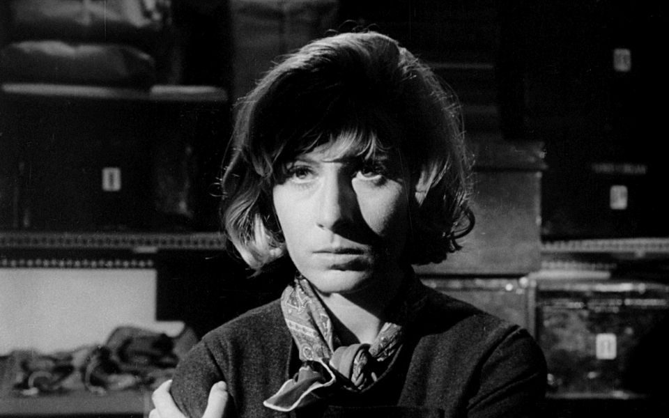 Black and white still. A woman with shoulder length hair and a neckerchief clutches her shoulder and looks into the distance.
