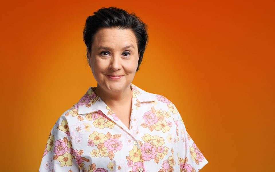 Susan Calman casually stands smiling, with hands in pockets, wearing a floral white shirt against an orange backdrop.