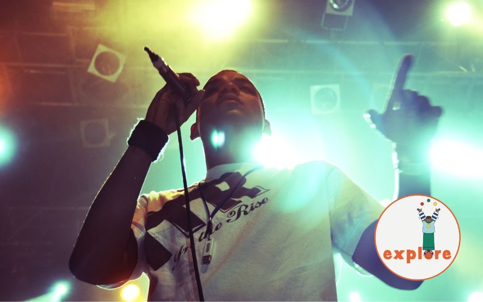Testament performs on stage in a t-shirt, holding a microphone in one hand and gesturing with the other, beneath fluorescent lights that flare on the camera. In the bottom right corner, a roundel featuring a playful illustration of a person juggling stood on the word 'explore'.