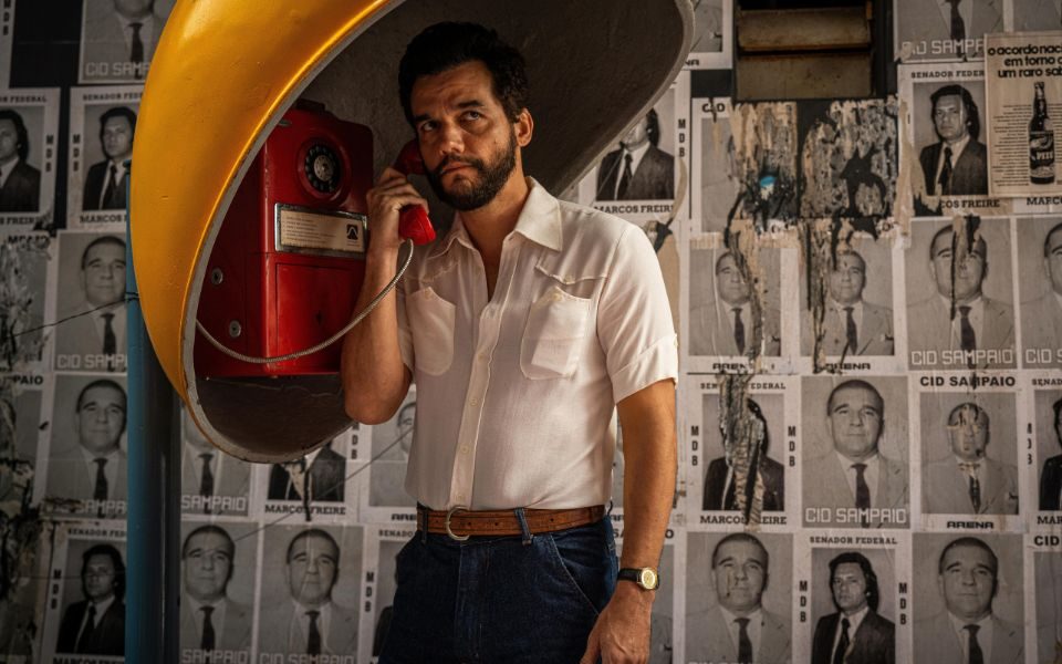 Wagner Moura stands at a vintage phone booth, the wall behind him covered in black and white posters of suited men.