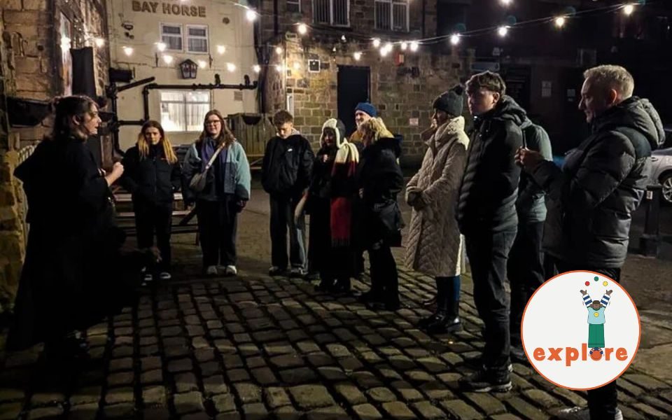 A caped person delivers a tour to a group of listeners, stood on a cobbled street at night. In the bottom right corner, a roundel featuring a playful illustration of a person juggling stood on the word 'explore'.
