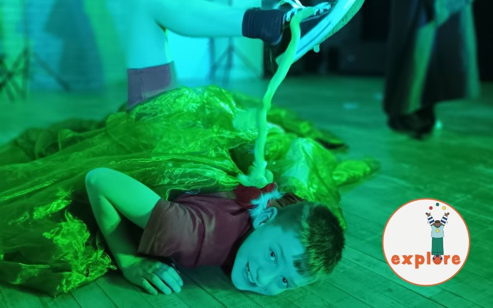 A boy is on the floor smiling up to the camera. He has green fabric draped across himself and is dangling a prop rose from his leg which is in the air. In the bottom right corner, a roundel featuring a playful illustration of a person juggling stood on the word 'explore'.