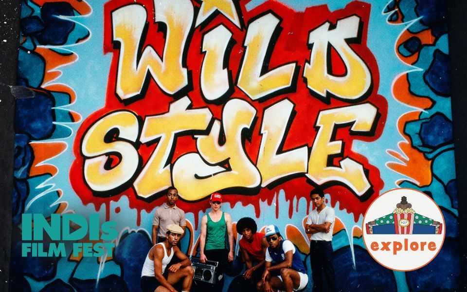 A group of teens dressed in 90s style outfits, baseball caps and trainers, one holding a boombox, crouch and pose in front of a graffitied wall, the text reading: Wild Style. Film Screening of the World First Hip-Hop Movie. In the bottom left, a turquoise logo reading 'INDIs Film Fest'. In the bottom right corner, a roundel featuring a playful illustration of a person sat peering over a bucket of popcorn, on the word 'explore'.