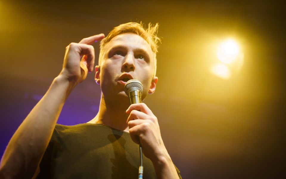 Lence speaking into a microphone, a stage light glowing above him.