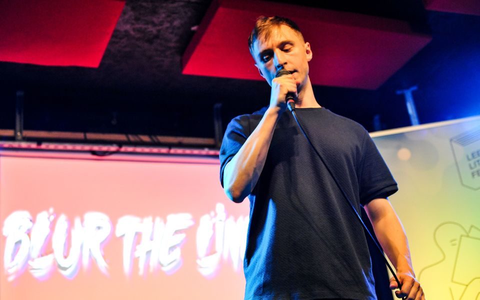 Lence on stage, speaking into a microphone. A projected slide behind him shows the logo for Blur the Lines.