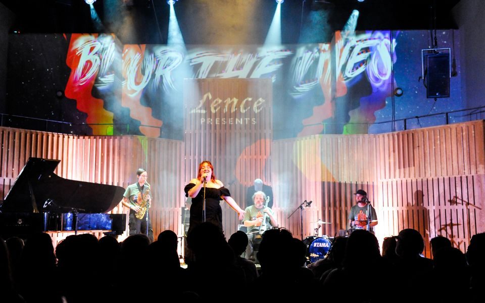 A singer stands centre stage at a microphone, a band playing behind her. Projections above her read Lence presents Blur the Lines