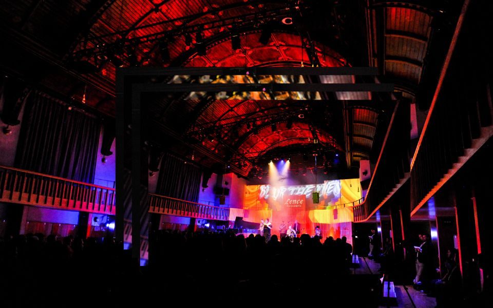 A wide shot of the interior of Howard Assembly Room, a full audience watching as a band performs beneath colour projections which read Lence presents Blur the Lines