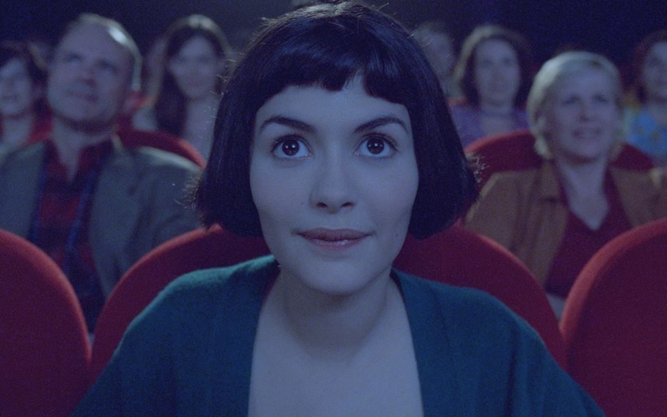 Amelie sits wide-eyed in a theatre amongst other viewers, looking towards a screen and lit by soft, blueish lighting.
