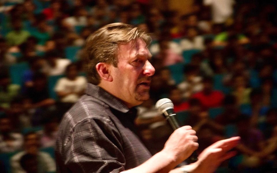 Andrew Newton performs to a packed blurry crowd, speaking into a handheld microphone and wearing a black shirt.
