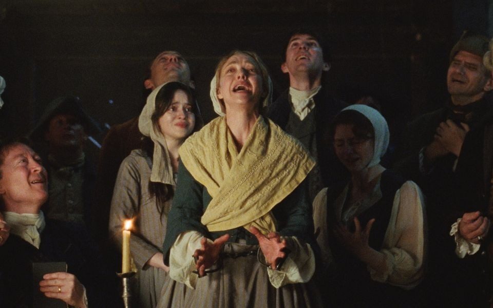 In a shawl and handcuffs, Amanda Seyfried passionately cries out amongst a group others in period clothing, clutching their chests.