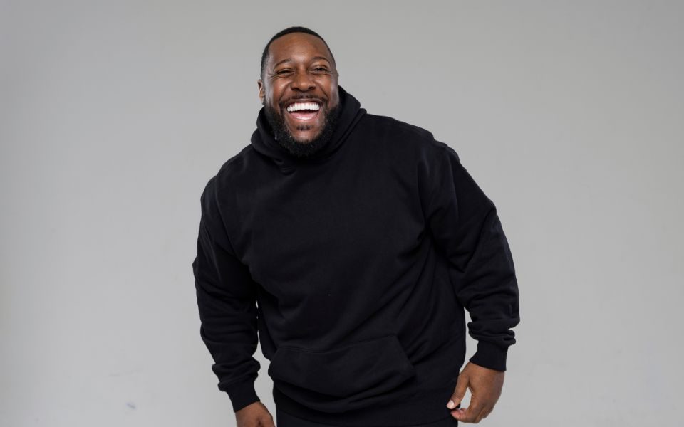 Aurie Styla is laughing and wearing a black hoodie, pictured against a grey backdrop.