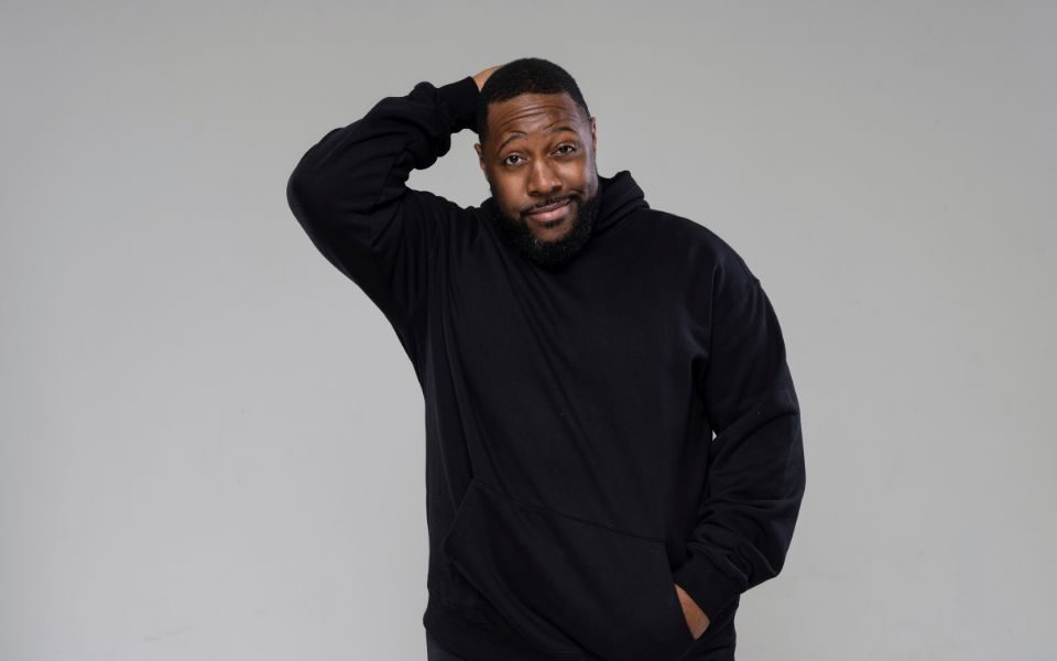 Aurie Styla smirks, with one hand held behind his head, the other in the pocket of his black hoodie, pictured against a grey backdrop.