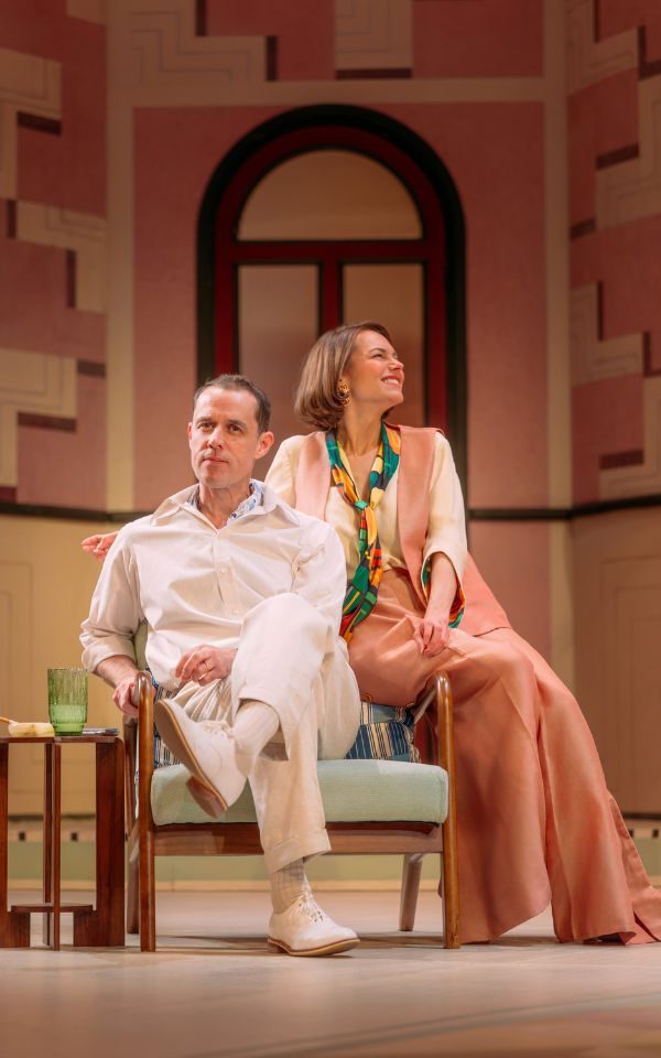 A woman wearing matching pink trousers and waistcoat perches on the arm of a chair with her arm around the man sitting in the chair, dressed in all-white.