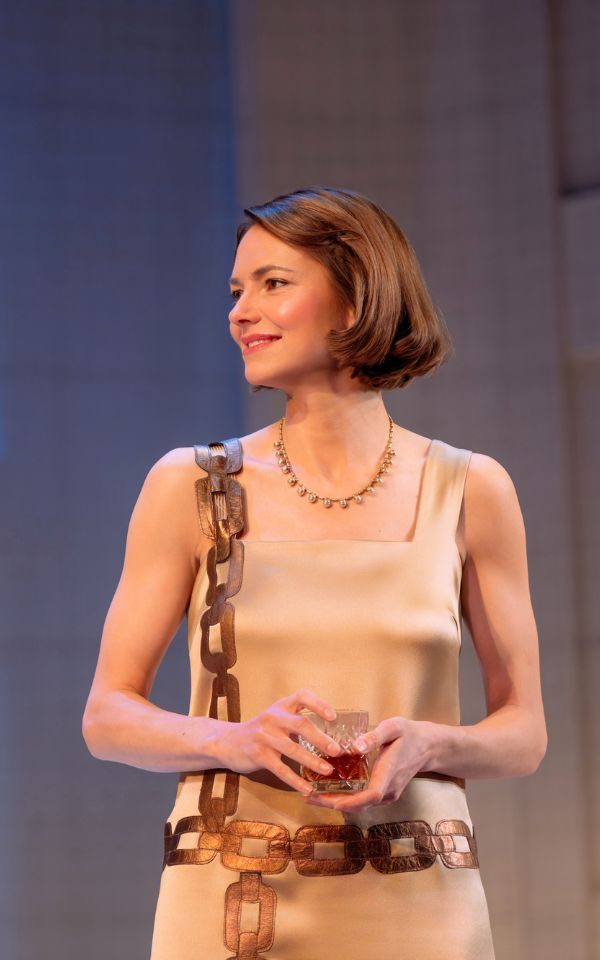 Kara Tointon as Constance in The Constant Wife. She wears a pale gold 1920s dress with a bronze chain pattern and holds a glass of whisky in her hand.