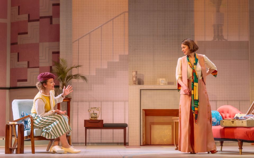 Two women in 1920s clothes talk to each other in a stark living room set, featuring a white wall with the shadow of a staircase, a fireplace and a small desk with a phone on it. One woman sits on a pale blue chair, wearing a green stripey skirt and yellow top with a purple baker-boy hat. The other woman stands up near a pink chaise-longue, wearing matching pale pink trousers and waistcoat with a green and yellow patterned scarf.