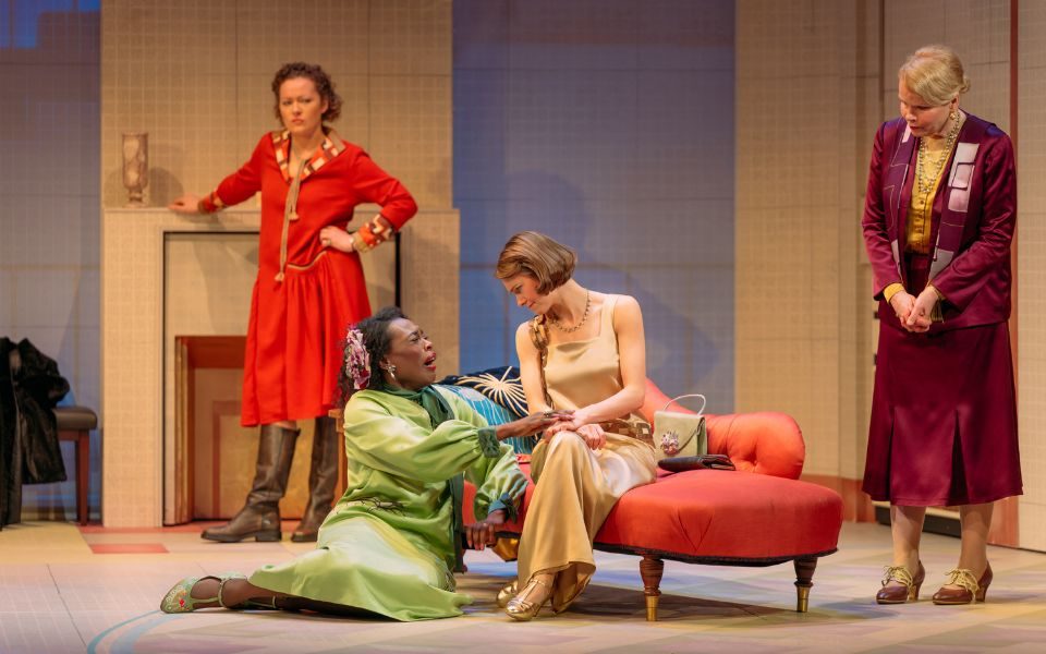 A woman in a green 1920s dress cries while sat on the floor, holding the hand of a woman in a pale gold dress who sits on an orange chaise-longue. Two other women stand around the room looking at them with concern.