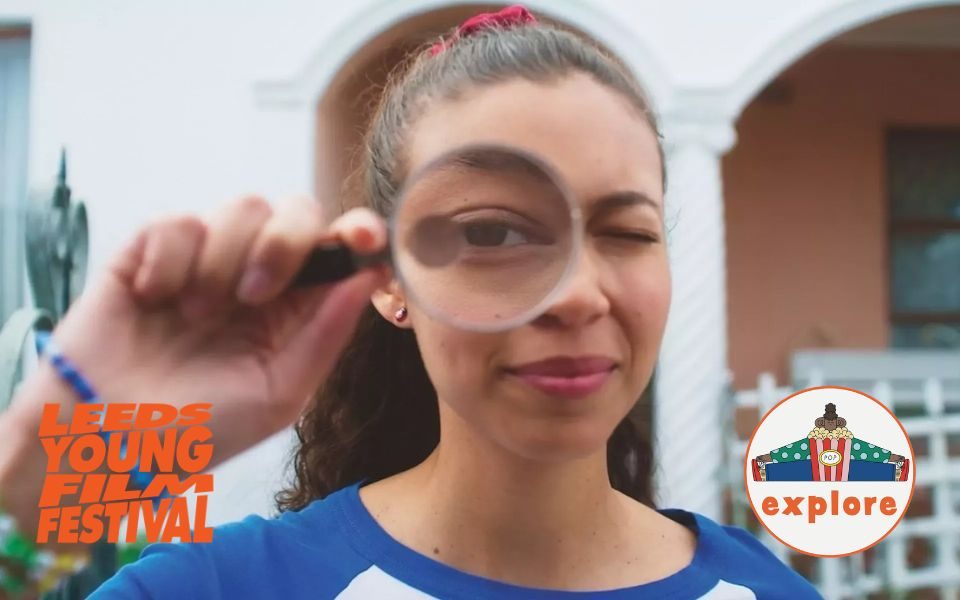 A girl looks into the camera lens through a magnifying glass. In the bottom left, an orange logo reading 'Leeds Young Film Festival'. In the bottom right corner, a roundel featuring a playful illustration of a person sat peering over a bucket of popcorn, on the word 'explore'.
