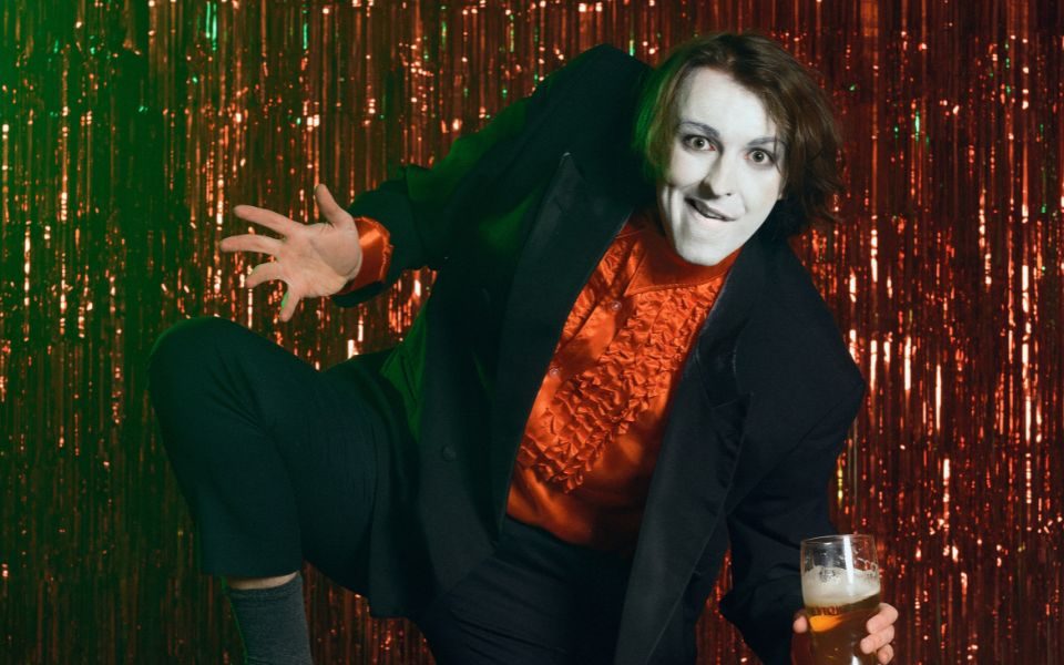 In white face paint and a frilly red shirt, Frankie Monroe grins and holds a pint of beer in front of a sparkly, red stage curtain.