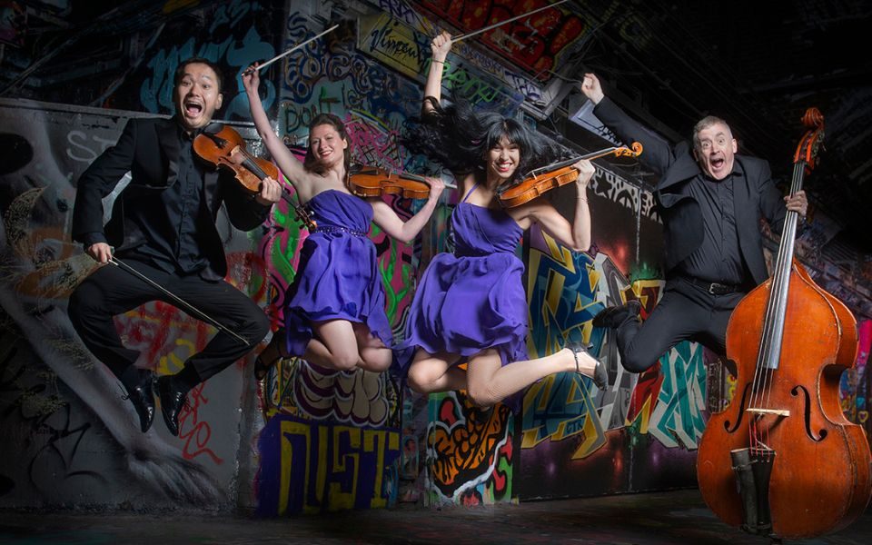 The quartet of Graffiti Classics jump up in the air with comic, open mouth expressions, each holding their instruments in a graffiti-covered tunnel.