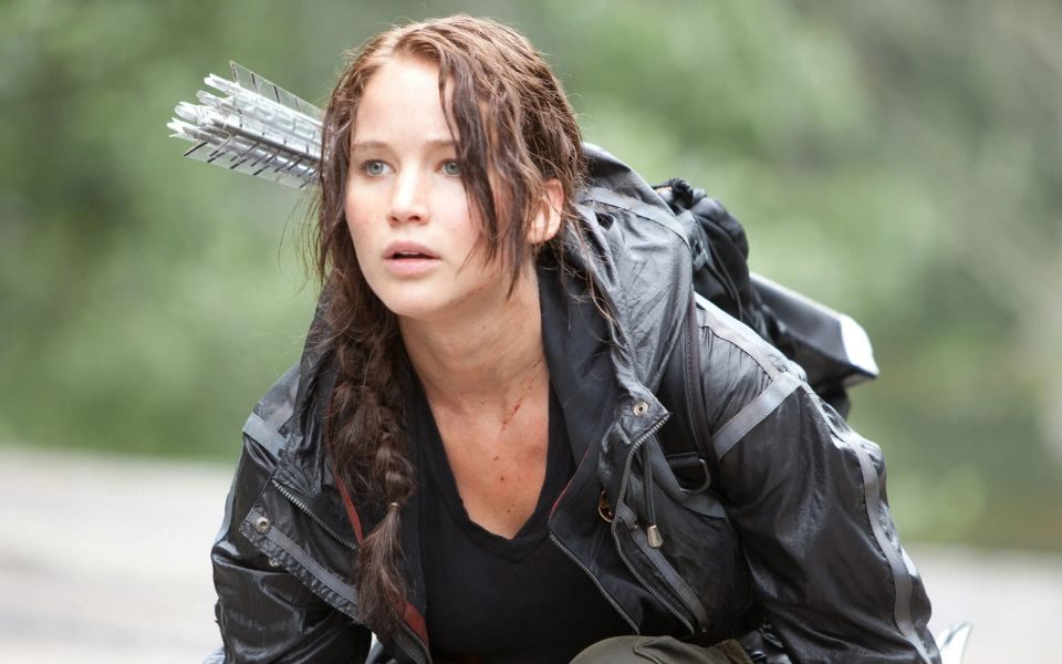 Jennifer Lawrence as Katniss in the Hunger Games, with plaited brown hair, black weatherproof coat and a quiver of arrows on her back.