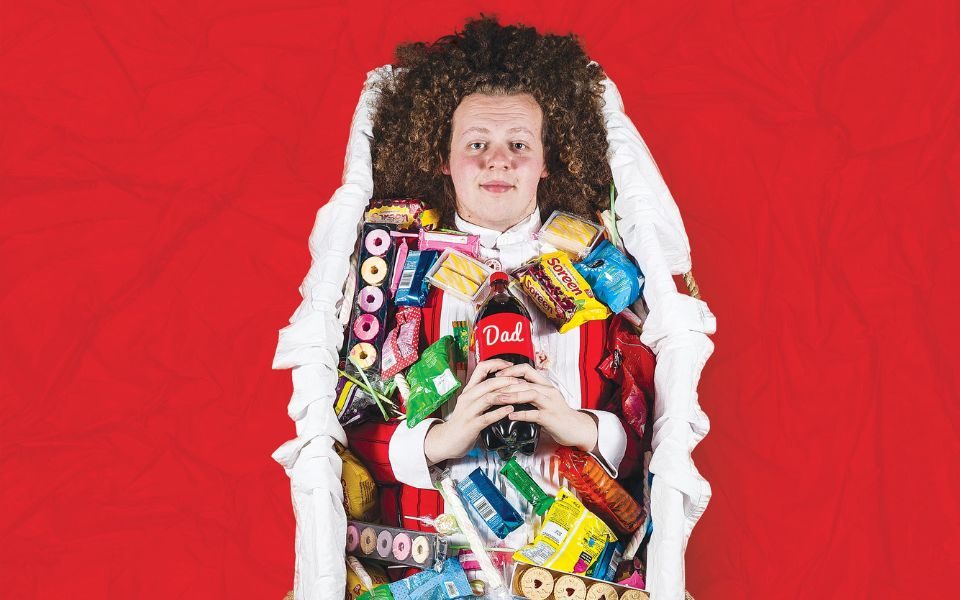 Jack Rooke lies in a coffin-shaped basket full of sweets and biscuits, holding a cola bottle with a label reading 'Dad', surrounded by red material.