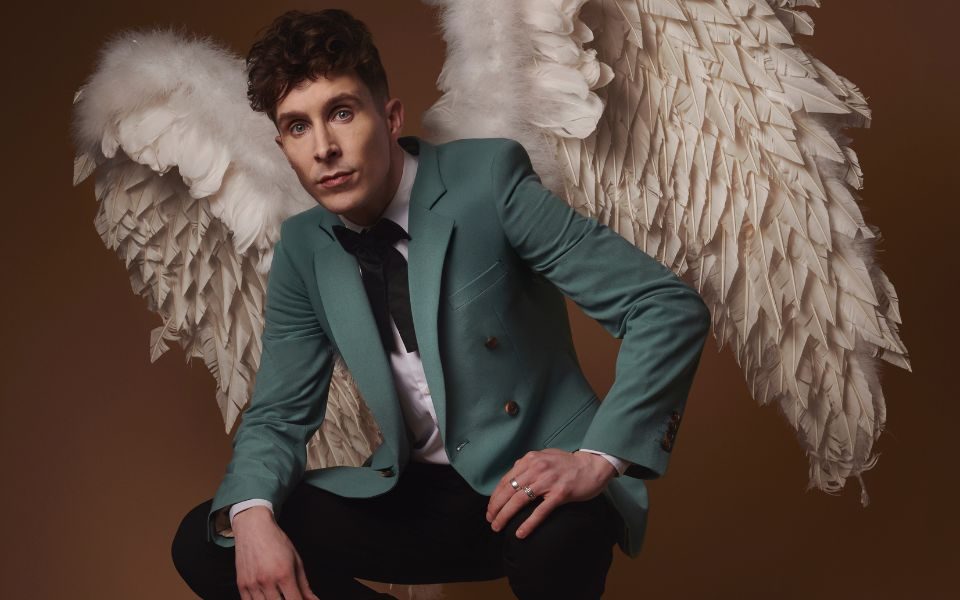 Larry Dean crouches in a suit with a turquoise jacket, resting his hands on his knees and wearing large angel wings on his back.