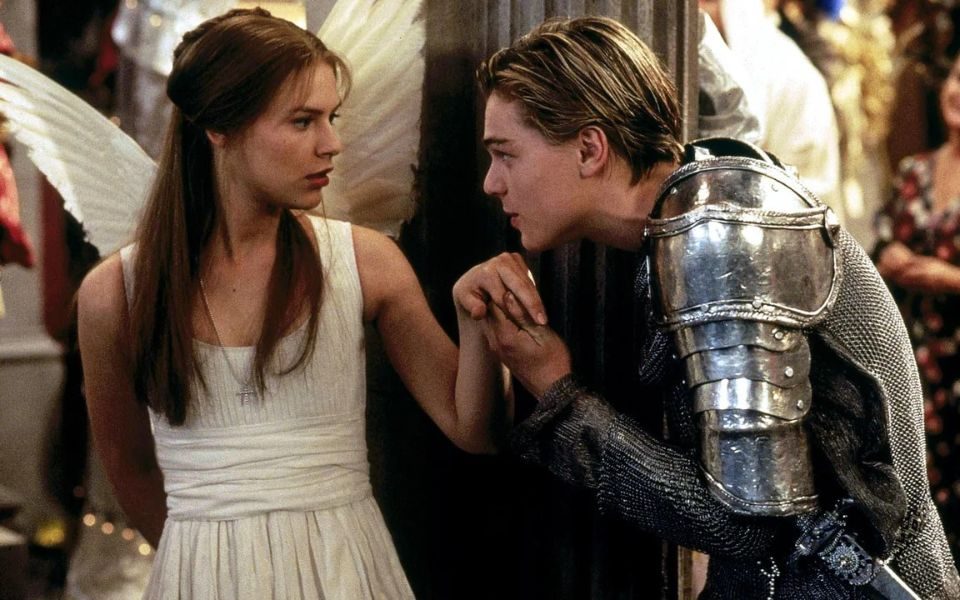 Leonardo DiCaprio with slicked back blonde hair and wearing a suit of armour holds the hand of Claire Danes, who is dressed in a white angel costume.