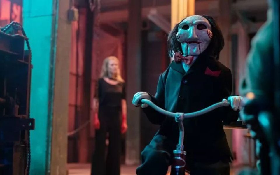 The puppet Jigsaw is on a tricycle, wearing a black suit with a red bowtie and face paint, lit by dramatic blue lighting. A woman with bloody hands is visible in the background.