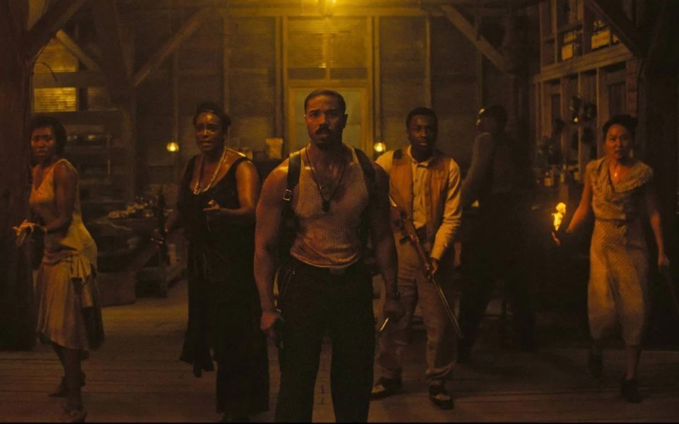 In a dimly lit barn, five characters in dirtied period clothing, led by Michael B Jordan, all carry weapons and stand together in defensive poses.