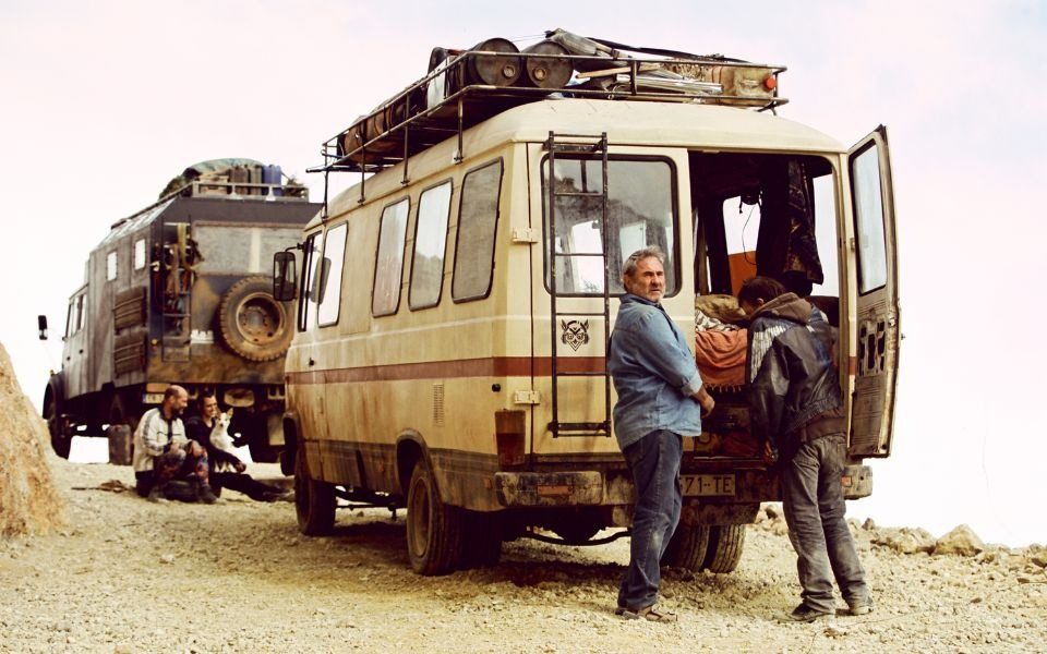 Two characters stand by the back of a rusty campervan in an arid desert setting.