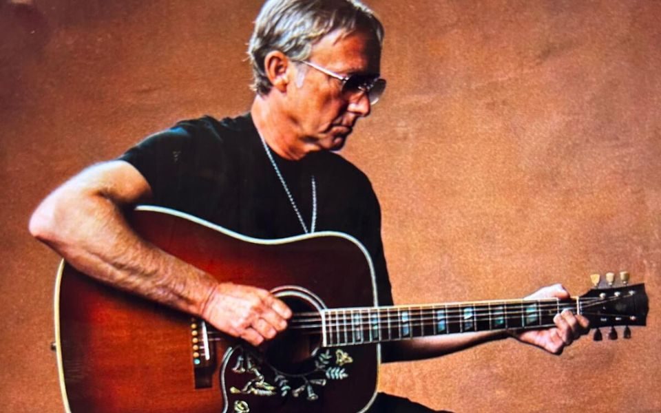 Steve Cradock sits on a stool and plays a patterned acoustic guitar, wearing a black t-shirt and sunglasses.