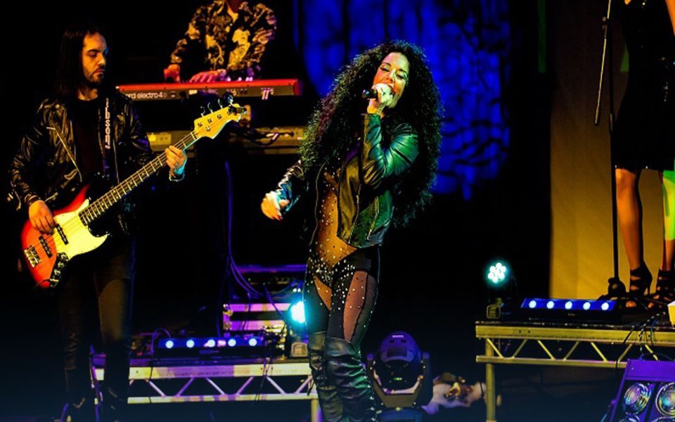 A Cher tribute performer sings into a handheld microphone beside a guitarist on stage. She has long, black, curly hair, leather jacket, knee-high boots and a glittery leotard.