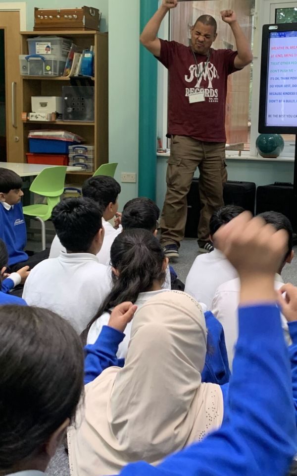 In a classroom, a crowd of children in school uniform raise their hands as they watch Testament at the front of the room. He wears a Jumbo Records t-shirt and has his eyes closed, fists raised above his head.