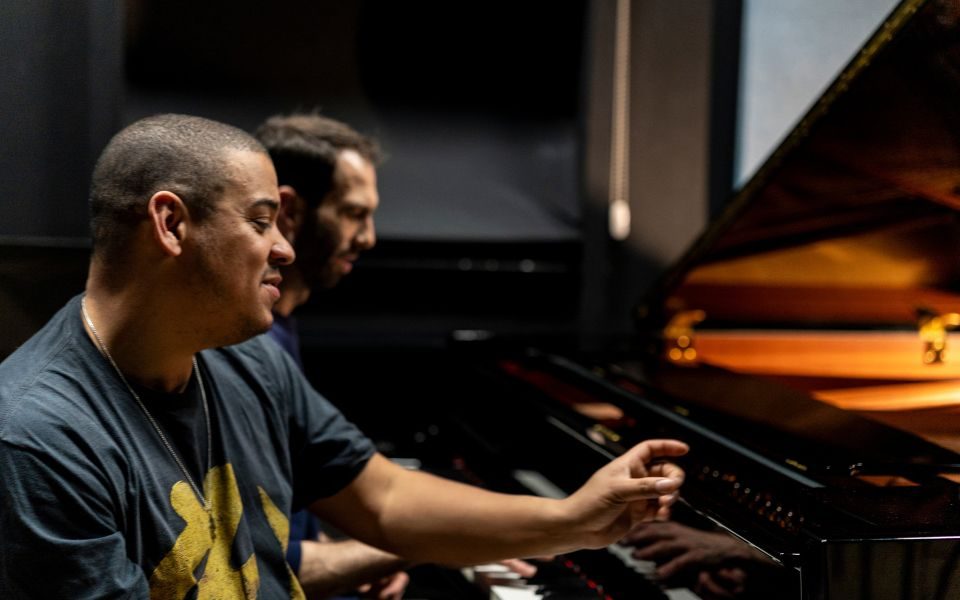 Testament smiles and gestures towards the keys of a grand piano, alongside Matthew Bourne who sits and plays the keys beside him.