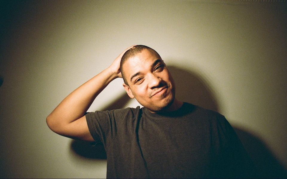 Testament smiles, shot under a spotlight against a blank backdrop, smirking and holding an arm behind his head.