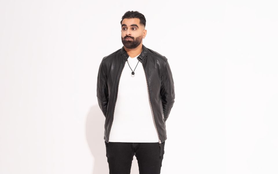 Tez Ilyas stands with his hands behind his back, wearing a leather jacket and white t-shirt as he looks off-camera in front of a white backdrop.