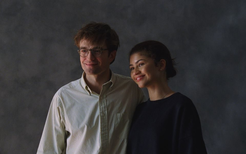 Robert Pattinson and Zendaya smile in a photographer's studio, against a mottled grey backdrop, posing for someone off-camera.