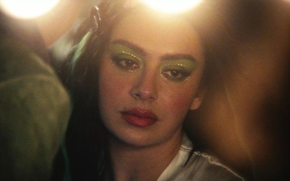 Charli XCX candidly glances off-camera, wearing green eye shadow embellished with gems, lit by hazy spotlights.