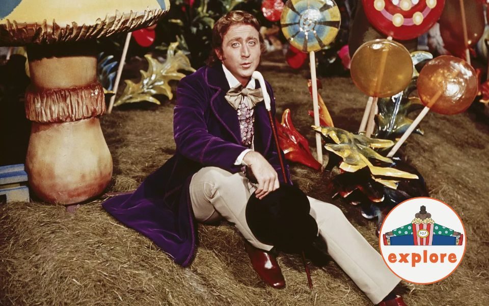 Gene Wilder as Willy Wonka, wearing a long purple jacket and extravagant bowtie, a cane resting on his torso. He sits wistfully on a bank beside large lollypops and sweets. In the bottom right corner, a roundel featuring a playful illustration of a person juggling stood on the word 'explore'.