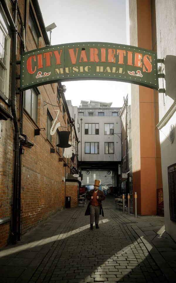A man in period costume, waistcoat and a large brown top hat walks down a sun-lit alleyway, beneath the arched sign for City Varieties Music Hall.