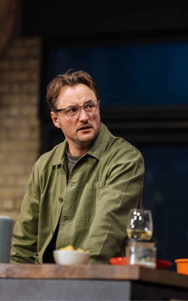 James Bye as Sam, wearing a khaki green shirt and glasses.