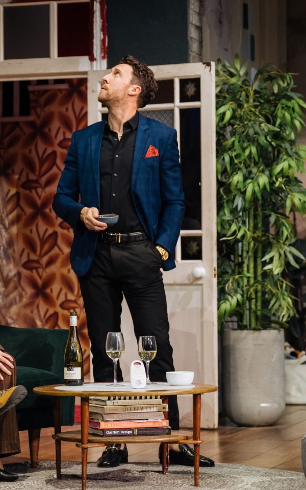 Grant Kilburn as Ben stands in a suit holding a small bowl and looking up at the ceiling.