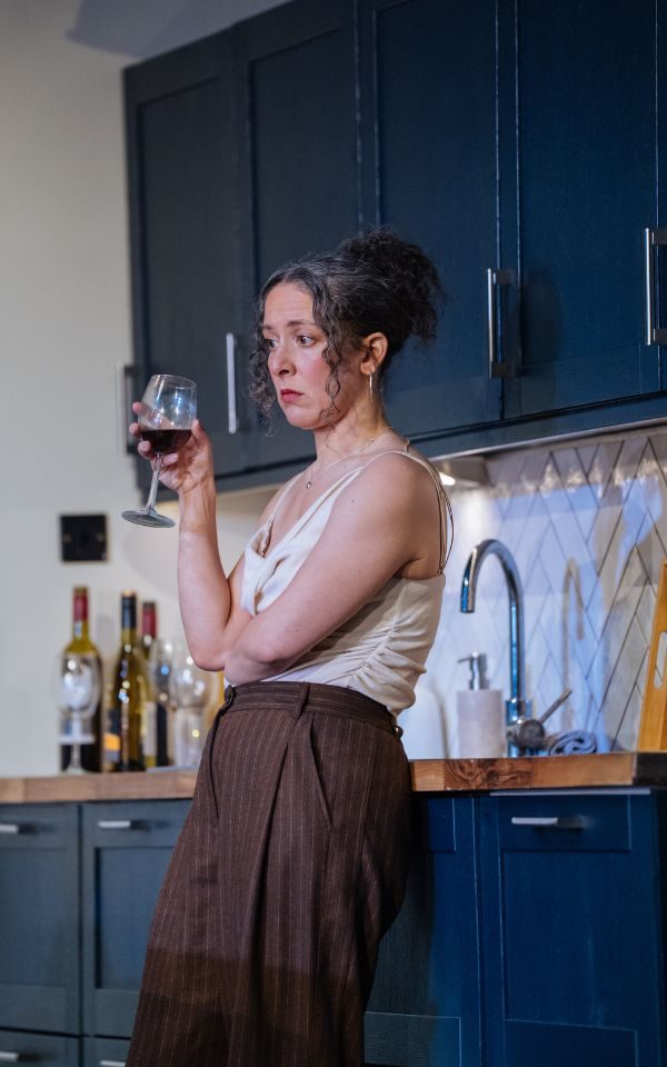 Natalie Casey as Lauren leaning against a kitchen cabinet with a glass of red wine in her hand.