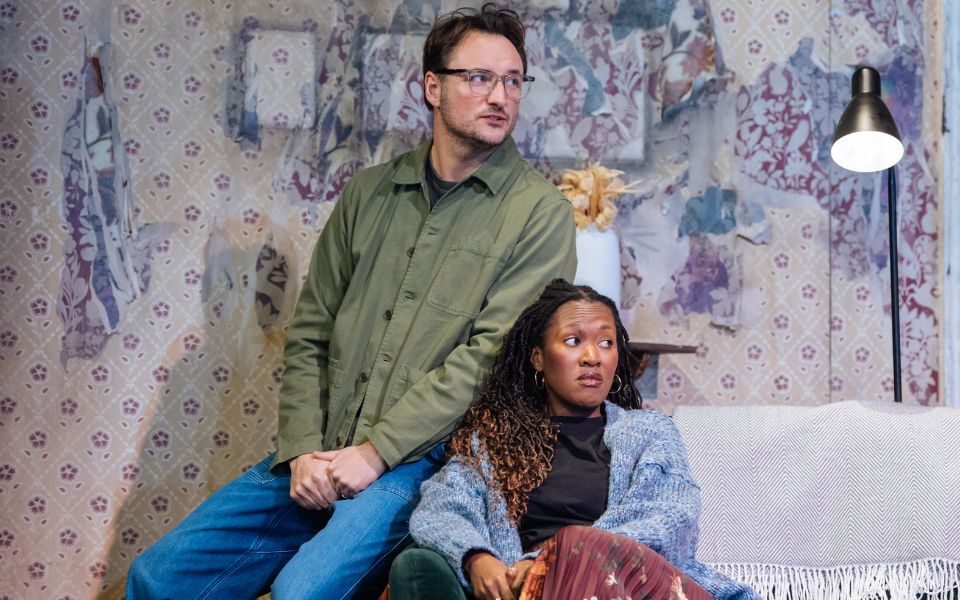 James Bye as Sam sitting on the arm of a sofa with Shvorne Marks as Jenny sat on the sofa seat beside him. Behind them is a shabby wall with torn wallpaper.