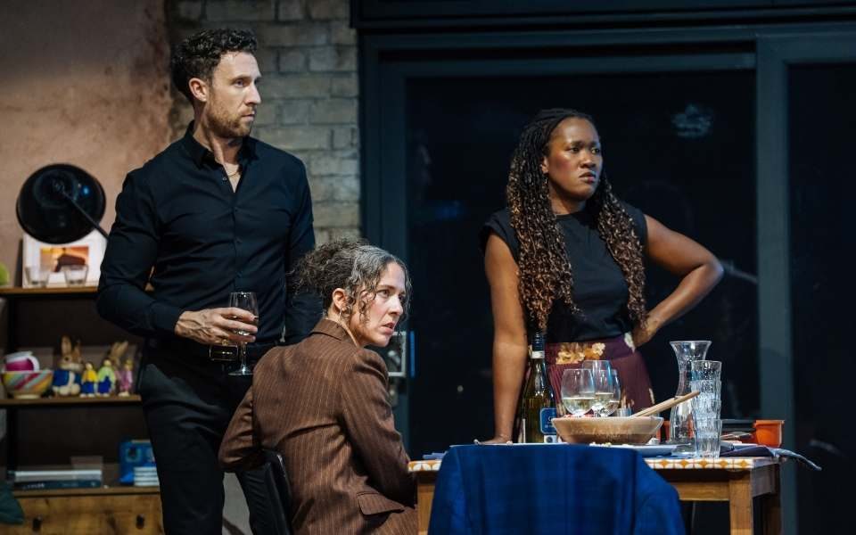 Natalie Casey as Lauren sat at the dinner table as Grant Kilburn as Ben and Shvorne Marks as Jenny stand around her.