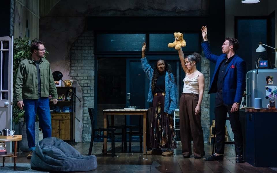 James Bye as Sam standing on one side of an open-plan living room/kitchen as Shvorne Marks as Jenny, Natalie Casey as Lauren and Grant Kilburn as Ben stand opposite him in a line, all raising one hand in the air. Natalie Casey holds up a stuffed bear.