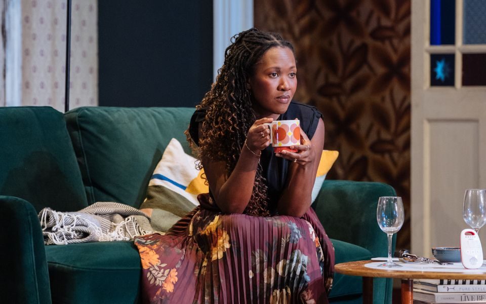 Shvorne Marks as Jenny sitting on the edge of a green sofa, holding a mug in her hands.